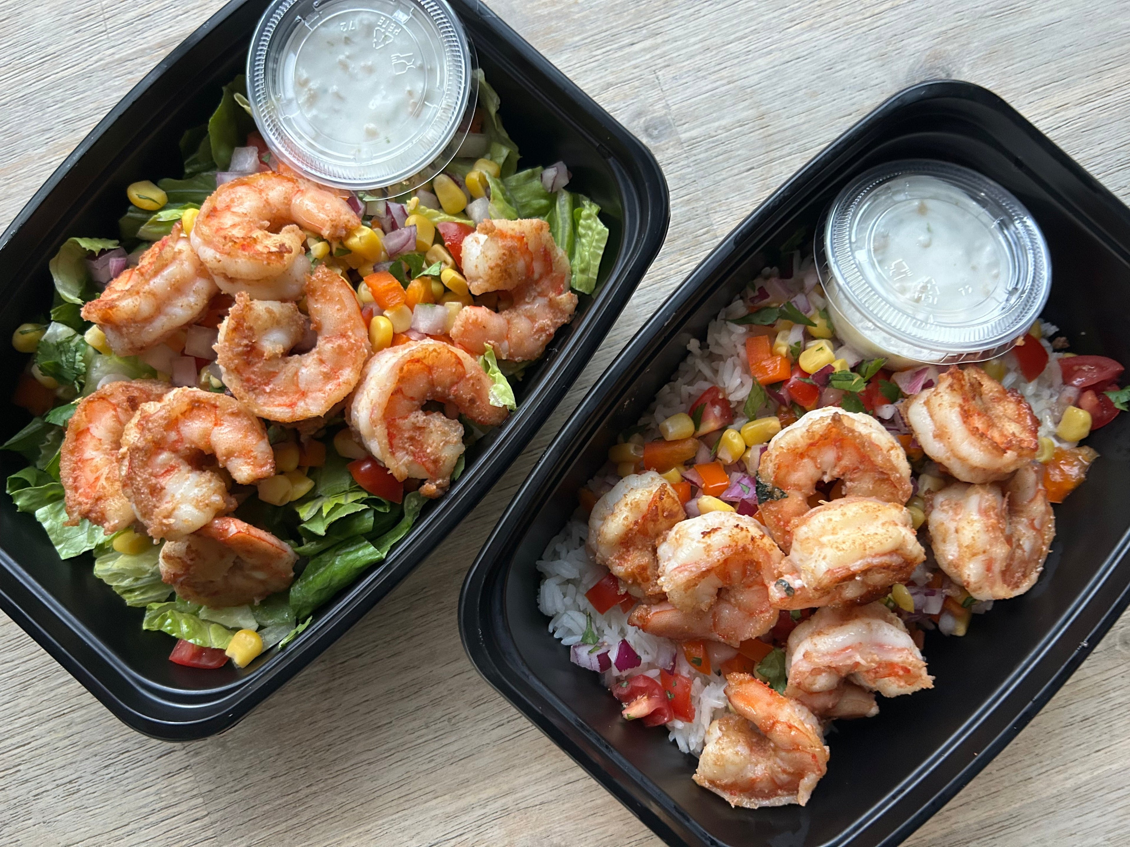 Two black meal prep containers with shrimp salads on a light wooden surface.